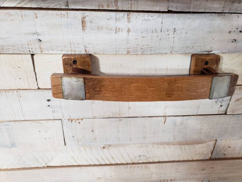 May include: A handcrafted wooden towel rack made from reclaimed oak barrel staves. The rack features a curved design with metal accents and is mounted on two rectangular wooden blocks. The wood has a natural, warm brown tone.