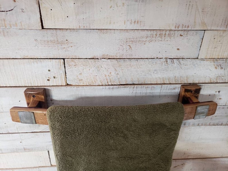 May include: A rustic wooden towel rack with a dark brown finish, mounted on a white-painted wooden wall. The rack features a dark green towel. The rack's design incorporates metal accents.