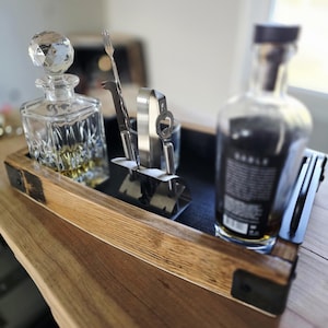 May include: A wooden serving tray with a black interior holds a crystal decanter, a bottle, and bar tools. The decanter has a faceted stopper. The tray has metal corner brackets and is on a wooden surface.