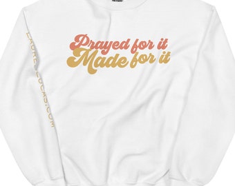 May include: White crewneck sweatshirt with the text "Prayed for it Made for it" in retro orange and gold lettering. The sleeve has the text "LAURELUCAS.COM" in gold. A casual, comfortable top.