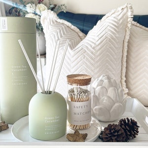 May include: A green tea and cucumber scented reed diffuser with a green glass bottle and a cork top. The diffuser is sitting on a white tray with a white ceramic artichoke, brown pine cones, and a jar of safety matches. The text on the bottle reads "Green Tea Cucumber Scented Reed Diffuser 120ml/4 fl oz CHICKIDEE".