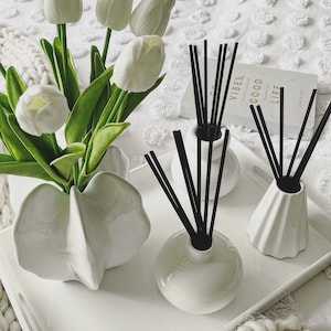 May include: A white ceramic vase containing white tulips alongside three white ceramic reed diffusers with black reeds arranged on a white rectangular tray. A book titled Good Vibes, Good Life is positioned behind the diffusers on the tray.