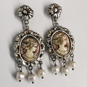 Puede incluir: Un par de pendientes de plata con un diseño de camafeo que representa el perfil de una mujer. Cada pendiente tiene un pequeño encanto de flor en la parte superior y tres gotas de perlas que cuelgan de la parte inferior.
