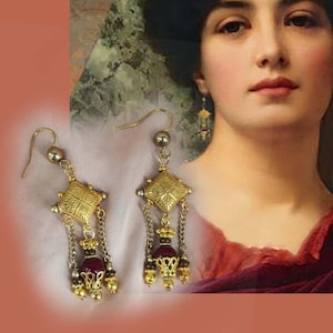 Op de afbeelding: Gouden oorhangers met een vierkante hanger, bordeauxrode kralen en gouden accenten.