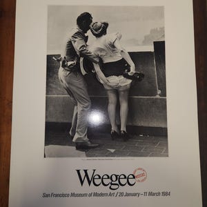 May include: Black and white photograph of a couple on a rooftop, with the woman's skirt blowing up in the wind. The photograph is a print from the San Francisco Museum of Modern Art, with the name "Weegee" and the dates "20 January-11 March 1984" printed below the image.
