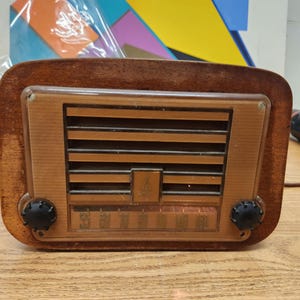 Puede incluir: Radio de madera vintage con marco rectangular y panel frontal marr&oacute;n claro. La radio presenta listones horizontales y un dial con n&uacute;meros. Dos perillas negras a cada lado. Una pieza cl&aacute;sica de decoraci&oacute;n del hogar.