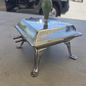 May include: A square, silver-toned metal kitchen appliance with a polished finish, featuring four curved legs, a central green handle on the lid, and two small metal prongs extending from one side of the base unit.