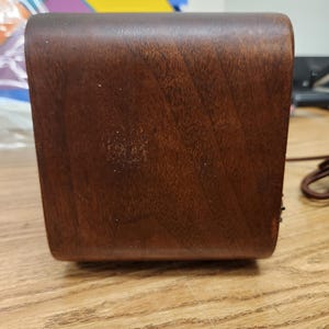 Puede incluir: Una caja rectangular de madera marr&oacute;n oscuro con bordes redondeados. La veta de la madera es visible y la caja parece estar bien hecha. Un cable marr&oacute;n es visible en el lado derecho. La caja est&aacute; sobre una superficie de madera marr&oacute;n claro.