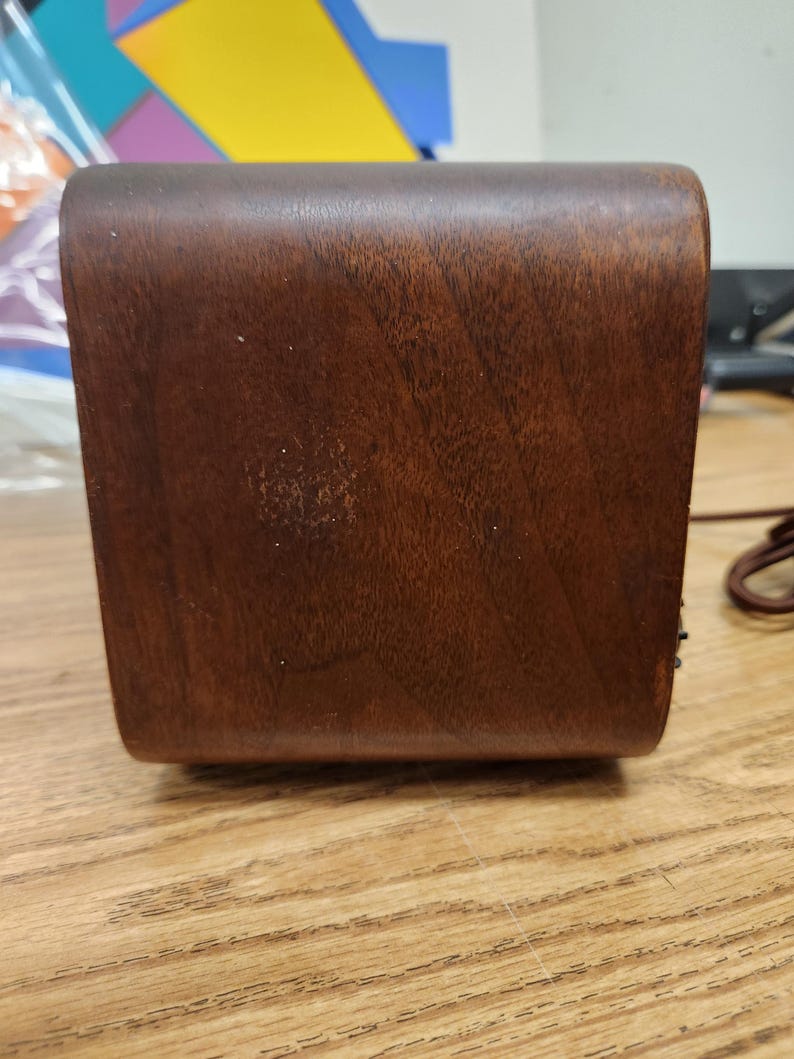 Puede incluir: Una caja rectangular de madera marr&oacute;n oscuro con bordes redondeados. La veta de la madera es visible y la caja parece estar bien hecha. Un cable marr&oacute;n es visible en el lado derecho. La caja est&aacute; sobre una superficie de madera marr&oacute;n claro.