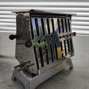May include: A vintage chrome-plated metal toaster featuring a rectangular body with vertical ventilation slots, a flat top, and a sturdy base. The appliance includes two black circular knobs on the side and two small metal prongs extending outward.