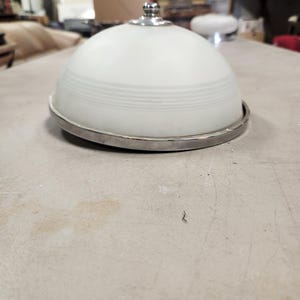 May include: A frosted glass light fixture with a silver-colored metal rim and a small decorative top. The fixture has a rounded, dome-like shape with horizontal etched lines. The light fixture is sitting on a light-colored surface.