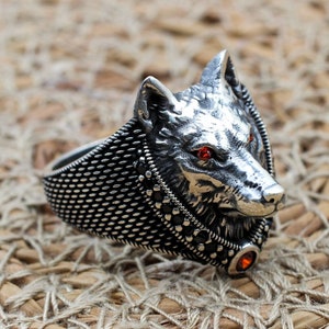 May include: A silver ring with a wolf head design. The wolf's eyes are red gemstones. The ring band is textured and has a small red gemstone on the side.