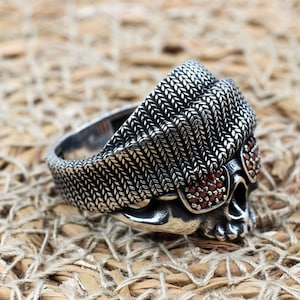 Puede incluir: Un anillo de calavera plateado con un diseño texturizado. La calavera presenta ojos de piedras preciosas rojas y un patrón tejido en la parte superior. El anillo se muestra sobre una superficie tejida de color canela.