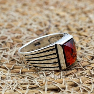 May include: A silver ring with a rectangular, faceted, red gemstone. The ring has a ribbed design on the band and is set against a woven, tan background. The ring is a piece of jewelry.
