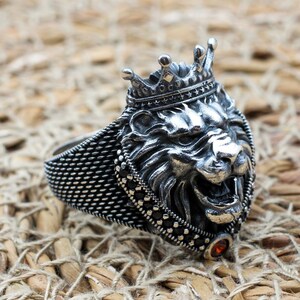 May include: A silver ring featuring a detailed lion's head wearing a crown. The ring has a textured band and is adorned with small, dark-colored stones. An orange-colored stone is set at the bottom of the lion's head.
