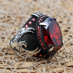 Può includere: Un anello d'argento con una grande pietra rossa e dettagli intricati. L'anello presenta un design a forma di uccello stilizzato ed è adornato con piccole pietre rosse.