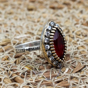 Op de afbeelding: Zilveren ring met een grote, ovale, dieprode edelsteen. De ring heeft een goudkleurige bezel rond de steen, geaccentueerd met kleine witte edelstenen. De band is zilver met een decoratief patroon. Op een geweven achtergrond.