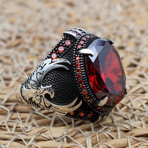 Può includere: Un anello d'argento con una grande pietra preziosa rosso scuro a taglio ovale come elemento centrale. L'anello presenta dettagli intricati, tra cui pietre rosse più piccole attorno alla gemma principale e motivi ornati sulla fascia. L'anello è posto su una superficie strutturata marrone chiaro.