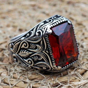 Könnte beinhalten: Ein silberner Ring mit einem verzierten Design und einem großen, roten Edelstein.