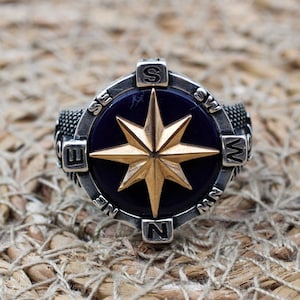 May include: A silver ring with a compass design. The compass face is blue with a gold star in the center. The ring is surrounded by a silver band with the letters N, S, E, and W.