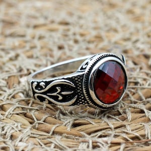 Op de afbeelding: Een zilveren ring met een ovale, gefacetteerde, rode edelsteen. De ring heeft een sierlijk ontwerp op de band en rond de edelsteen. De achtergrond is een getextureerd, neutraal getinte ondergrond.