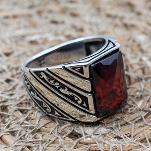 May include: A silver ring with a large, rectangular, deep red gemstone. The ring has a geometric design with black accents on the sides. The ring is set against a textured, woven background.
