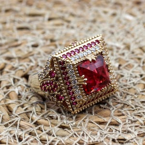May include: A gold-coloured ring featuring a large, square, red gemstone. The ring is embellished with smaller red and white gemstones, set against a woven, beige background. The ring's design is intricate and detailed.
