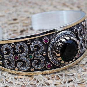 May include: A silver bracelet with intricate black and gold detailing. The bracelet features a large, round, black gemstone surrounded by smaller white gemstones. The bracelet is decorated with small red and green gemstones.