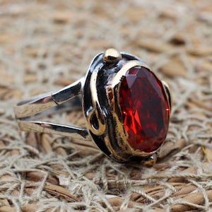 May include: A silver ring with a red gemstone set in a gold-toned setting. The ring has a double band and a decorative design.