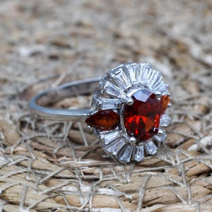 May include: A silver ring with a large red oval gemstone surrounded by smaller clear gemstones.
