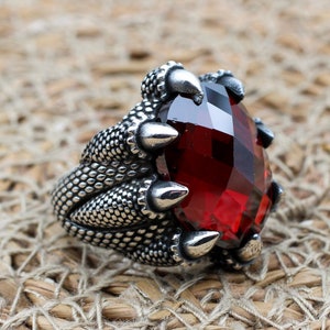 Peut inclure: Une bague en argent avec une pierre précieuse rouge sertie dans un design en griffes. La bague est détaillée avec de l'argent texturé.