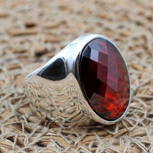 Può includere: Un anello d'argento con una grande gemma rosso scuro, tagliata a forma ovale e sfaccettata. L'anello ha una superficie lucida e riflettente, su uno sfondo tessuto strutturato. Le sfaccettature della gemma brillano.