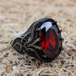 Peut inclure: Une bague en argent ornée d'une grosse pierre précieuse rouge foncé de forme ovale. La monture de la bague présente des détails noirs complexes autour de la pierre précieuse et sur les côtés. La bague est posée sur un fond texturé et tissé.