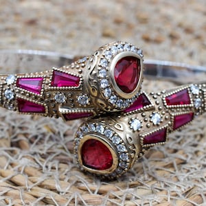 May include: A silver bangle bracelet with a decorative design featuring red and clear gemstones. The bracelet is open and has a hinged clasp.