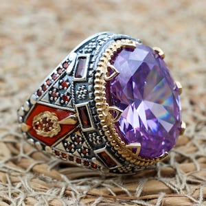Peut inclure: Une bague en argent avec une grande pierre précieuse violette entourée de petites pierres précieuses rouges. La bague présente des détails complexes et une bande dorée.