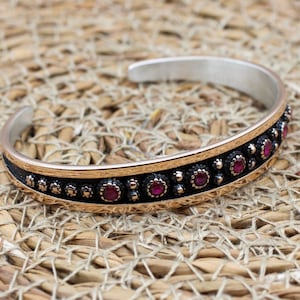 Puede incluir: Brazalete de puño plateado y dorado con una banda negra. El brazalete presenta una fila de pequeñas piedras preciosas rojas redondas y un patrón decorativo. El brazalete se muestra sobre una superficie tejida de color canela.