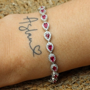 May include: A silver bracelet featuring pear-shaped red gemstones and small clear stones. The bracelet is worn on a wrist with a tattoo that says "Aslan" with a heart symbol. The background is a woven, tan surface.