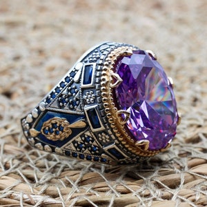 Könnte beinhalten: Ein silberner Ring mit einem großen, facettierten, violetten Edelstein, der in einer goldfarbenen Fassung sitzt. Der Ring ist mit komplizierten Details und blauen Steinen verziert.