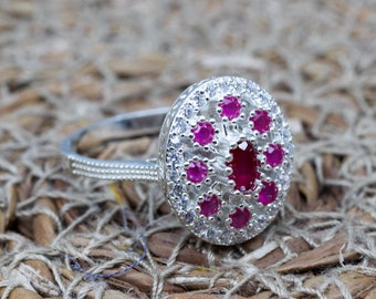 May include: A silver ring with a cluster of small, round, bright pink gemstones surrounding a larger oval pink gemstone. The ring is set against a textured, woven background.