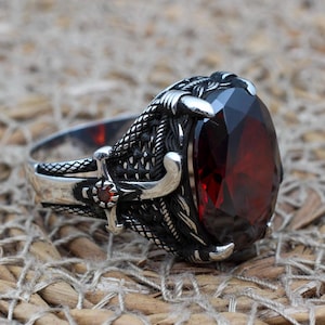 Könnte beinhalten: Ein silberner Ring mit einem großen, dunkelroten Edelstein, der in einem detaillierten, klauenartigen Design gefasst ist. Der Ring liegt auf einem gewebten, braunen Untergrund.