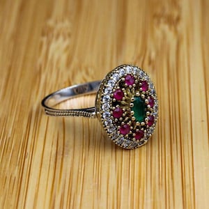 May include: A silver ring featuring an oval-shaped centrepiece with a central green gemstone surrounded by a halo of small red stones and an outer border of clear white stones. The band has a textured, rope-like pattern along the sides.