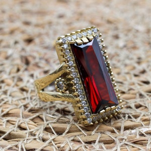 Puede incluir: Anillo dorado con una gran gema rectangular de color rojo oscuro como pieza central. La gema está rodeada por un borde de piedras más pequeñas y transparentes. La banda del anillo presenta detalles intrincados y piedras pequeñas adicionales.