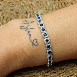 May include: A silver tennis bracelet with blue square-shaped stones set in a halo of smaller stones. The bracelet is on a person's wrist.