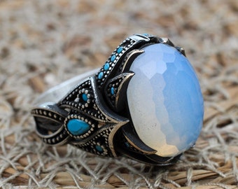May include: A silver ring featuring a large, faceted, oval, pale blue gemstone. The ring has intricate detailing with small turquoise stones and a decorative band. The ring is set against a textured, woven background.