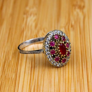 May include: An oval-shaped ring featuring a central red stone surrounded by smaller red stones and a border of clear white stones. The setting is a dark, textured metal with gold-toned accents, and the band has a detailed, etched pattern along the sides.