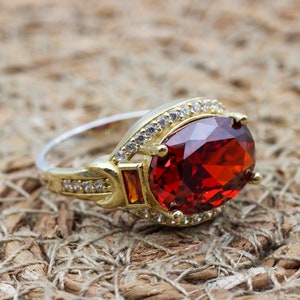 Puede incluir: Un anillo de oro y plata con una gran piedra preciosa roja oscura de talla ovalada como pieza central. El anillo está adornado con piedras preciosas más pequeñas y transparentes y una banda dorada. El anillo está sobre un fondo texturizado y tejido.