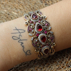 Puede incluir: Un brazalete de oro con intrincados diseños de filigrana y piedras preciosas rojas y transparentes. El brazalete está en la muñeca de una persona con un tatuaje que dice "Aslam".