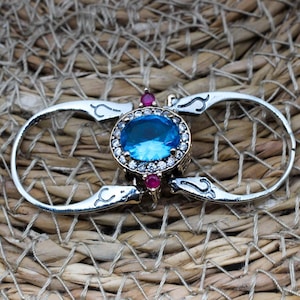 Peut inclure: Une bague en argent avec une grande pierre précieuse bleue entourée de petites pierres blanches. La bague a un design décoratif avec deux boucles de chaque côté de la pierre précieuse. La bague est posée sur un fond tissé.