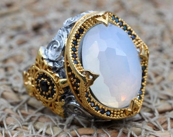 May include: An elaborate ring with a large, oval, pale blue gemstone. The ring has a gold-coloured setting with detailed patterns and small black accents. The band is silver-coloured with gold embellishments.