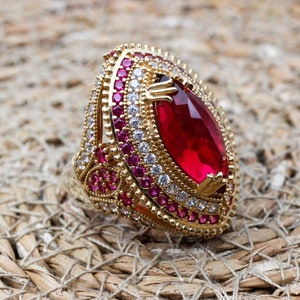 Könnte beinhalten: Ein goldfarbener Ring mit einem großen, tropfenförmigen, roten Edelstein. Der Ring ist mit kleineren roten und weißen Edelsteinen besetzt, die ein detailliertes Muster bilden. Präsentiert auf einer gewebten, beigefarbenen Oberfläche.